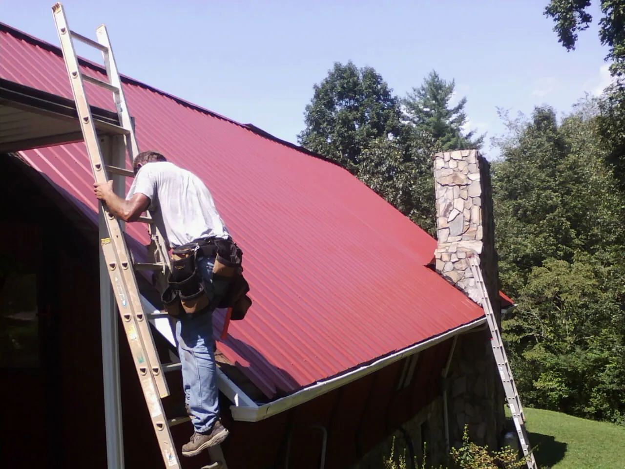 Roofer on a ladder beside a red metal roof and stone chimney — Soffit & Fascia Repair by Roofing Brothers in Walthall